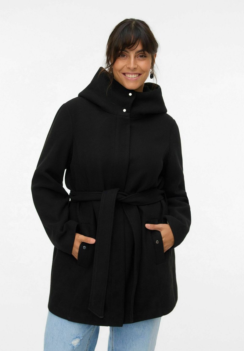 Manteau noir à capuche avec une ceinture, fabriqué en tissu doux. Il présente un col à pression, deux poches avant et est associé à un jean bleu clair.