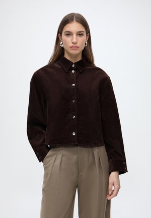 Femme portant une chemise en velours côtelé marron foncé boutonnée et un pantalon plissé beige sur un fond clair uni.