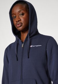 Marineblå hettegenser med glidelås laget av mykt stoff, med kengurulomme, snorhette og brodert Champion-logo på brystet.