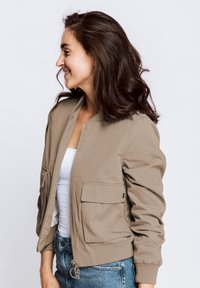Khaki bomber jacket with zip closure, two front pockets, ribbed cuffs, and a smooth texture. Worn over a white top and blue jeans.