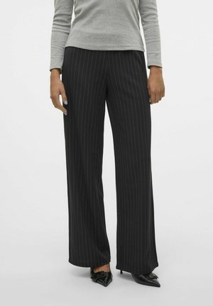 Osoba w szerokich, czarnych spodniach w prążki, szarym topie z długim rękawem i czarnych szpilkach ze spiczastym noskiem stoi na jednolitym tle.