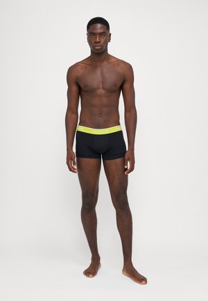 Schwarze Boxershorts mit einem limettengrünen Lacoste-Logo auf dem Bund. Der Stoff wirkt glatt und körpernah, was ein modernes Design zur Schau stellt.