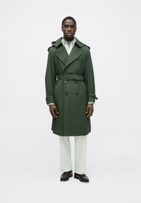 Trench verde con bottoni doppiopetto, cintura e forma accentuata da spalline alle spalle, abbinato a pantaloni chiari e scarpe.