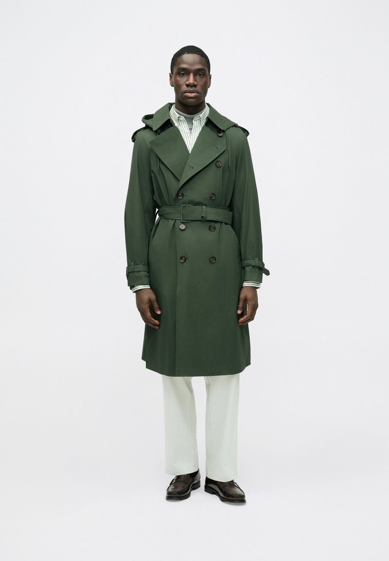 Trench verde con bottoni doppiopetto, cintura e forma accentuata da spalline alle spalle, abbinato a pantaloni chiari e scarpe.