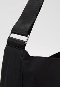 Schwarze Stoff-Schultertasche mit einem Textilgurt, ausgestattet mit einer silbernen Metallringbefestigung und einer glatten, strukturierten Oberfläche.