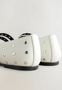 Bílé ploché boty vyrobené z hladkého syntetického materiálu, se stříbrnými kovovými hřebíčky, kulatou špičkou a černým vnitřním lemováním s texturovanou podrážkou.