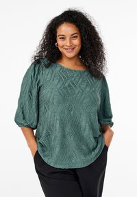 Textured green blouse with a wavy pattern, round neckline, puff sleeves, and a relaxed fit. Paired with black trousers.