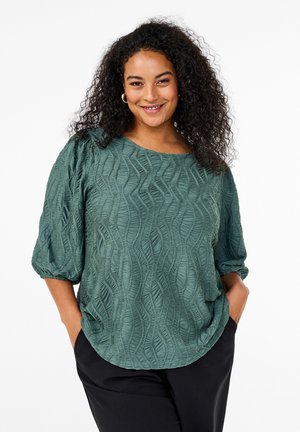 Textured green blouse with a wavy pattern, round neckline, puff sleeves, and a relaxed fit. Paired with black trousers.