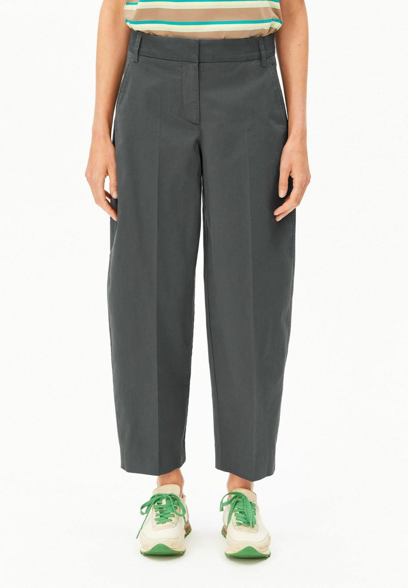 Person, die dunkelgraue, gekürzte Hose, ein gestreiftes Hemd und weiße Turnschuhe mit grünen Schnürsenkeln trägt, steht vor einem weißen Hintergrund.