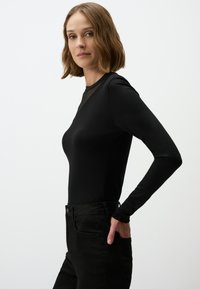 Body nero a maniche lunghe con texture liscia e dettaglio trasparente sul collo, abbinato a jeans neri a vita alta. Il modello posa di lato.