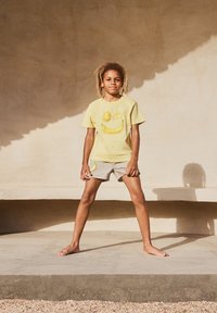 T-shirt gialla con faccina sorridente e illustrazione di frutta, abbinata a pantaloni corti beige con design di banana; indossata a piedi nudi.