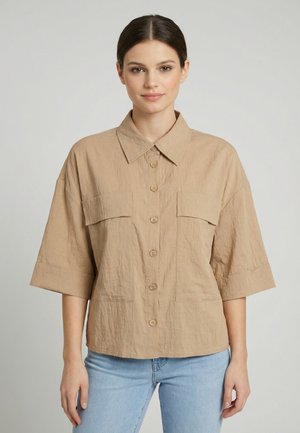 Femme portant une chemise beige ample à boutons avec de larges manches et deux poches avant, associée à un jean bleu clair, debout devant un fond uni.
