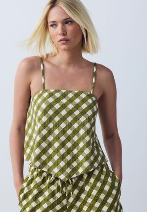 Donna bionda con top senza maniche a quadri verdi e bianchi e pantaloni coordinati con coulisse, che guarda leggermente di lato con le mani nelle tasche.