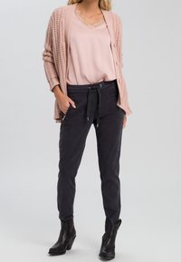Cardigan en tricot rose clair, superposé à un blouse rose avec des bordures en dentelle, associé à un pantalon noir fuselé et des bottes à talons noires.