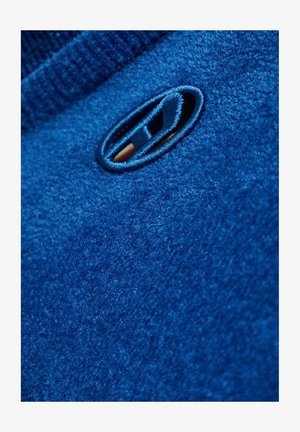 Indumento in pile blu con una texture liscia, scollo a coste e un dettaglio cucito prominente a forma ovale vicino al colletto.