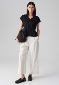 T-shirt nera a scollo a V, vestibilità ampia, indossata con pantaloni larghi color crema. Il modello tiene una borsa nera. Mocassini neri completano l'outfit.