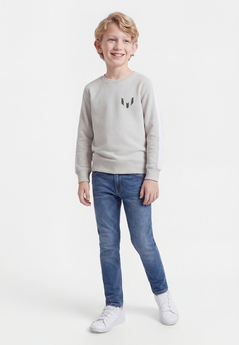 Ενα χαμογελαστό αγόρι που φοράει ανοιχτό γκρι μακρυμάνικο μπλουζάκι με λογότυπο, μπλε τζιν και λευκά αθλητικά παπούτσια, στέκεται μπροστά σε απλό λευκό φόντο.