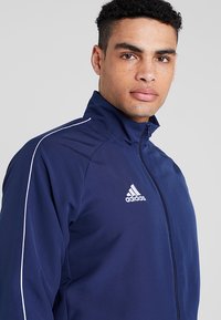 Marineblaue Sportjacke mit hohem Kragen, Frontreißverschluss und weißen Streifen an den Ärmeln. Mit aufgesticktem weißen Adidas-Logo auf der Brust.