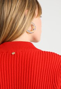 Roter gerippter Pullover mit hohem Kragen und einem metallischen Logostick auf dem Rücken; kombiniert mit großen, gemusterten Creolen.