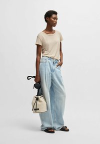T-shirt beige a maniche corte, jeans ampi blu chiaro, sandali neri e una borsa a secchiello crema con logo "BOSS" e dettagli neri.