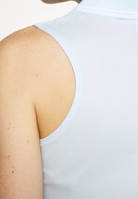 Närbild av en persons axel och överarm som bär en ljusblå ärmlös skjorta med krage.