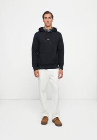 Sudadera negra con bolsillo frontal y logo "GANT" en blanco, combinada con pantalones blancos y zapatos de ante marrones, de pie frente a un fondo blanco.