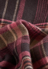 Woolen fabric featuring a plaid pattern with dark brown, pink, and tan colors. Soft texture with visible fibers and stitched edges.