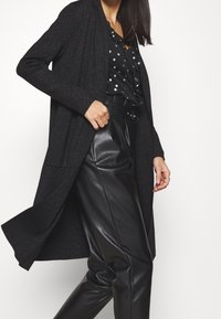 Svart lång cardigan med texturerad yta, bärs över en svart blus med prickar, tillsammans med högmidjade svarta läderbyxor.