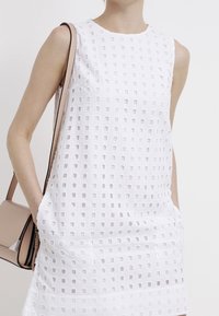 Robe blanche sans manches avec un motif texturé de petites découpes carrées. Comprend des poches latérales et un col rond.