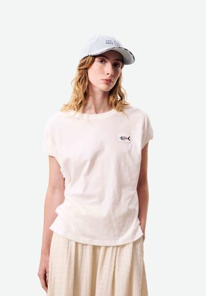 Jeune femme aux cheveux blonds ondulés porte un t-shirt blanc avec un petit écusson en forme de poisson et une jupe claire, posant devant un fond clair uni.