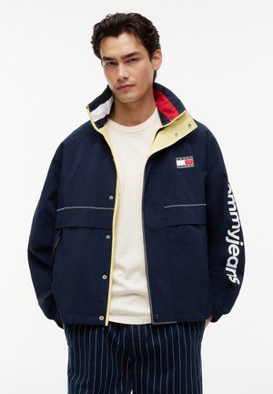 Ung mand iført en marineblå jakke med Tommy Jeans-logo på ærmet og brystet, cremefarvet skjorte og marineblå bukser med dertil hørende striber, stående mod hvid baggrund.