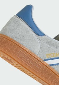 Adidas sporta apavi ar pelēku zamšādu augšdaļu, zilas ādas akcentu, baltu svītru un oranžu gumijas zoli ar teksturētu rakstu.