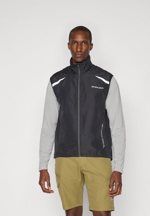 Veste sans manches noire avec un col haut, des accents blancs et des détails texturés aux épaules, portée sur une chemise à manches longues gris clair et un short beige.