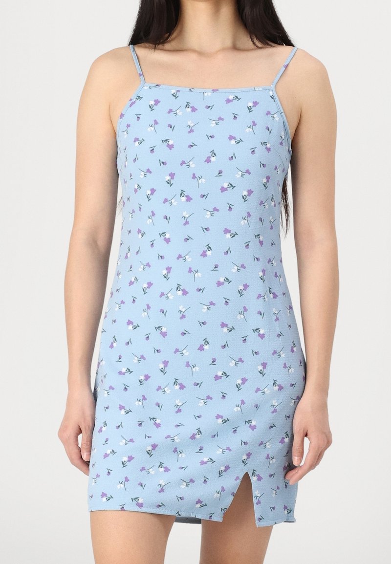 Robe mini bleue sans manches avec fines bretelles, petit imprimé floral violet et blanc, et fente avant au-dessus du genou, portée par une personne debout.