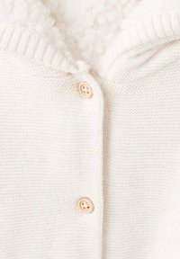 Vit stickad cardigan med en mjuk, texturerad insida, en ribbad krage och två träliknande knappar för stängning.