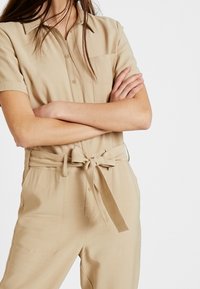 Jumpsuit beige à manches courtes avec un devant boutonné, coupe ajustée, une poche poitrine et une taille ceinturée mettant en valeur un tissu léger et lisse.