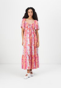 Kvinde stående i en pink blomstret midi-kjole med flagrende ærmer og hvide sko mod en ensfarvet hvid baggrund.