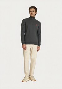 Joven vestido con un suéter gris oscuro con cremallera, pantalones beige claros y zapatillas beige, de pie frente a un fondo sencillo.