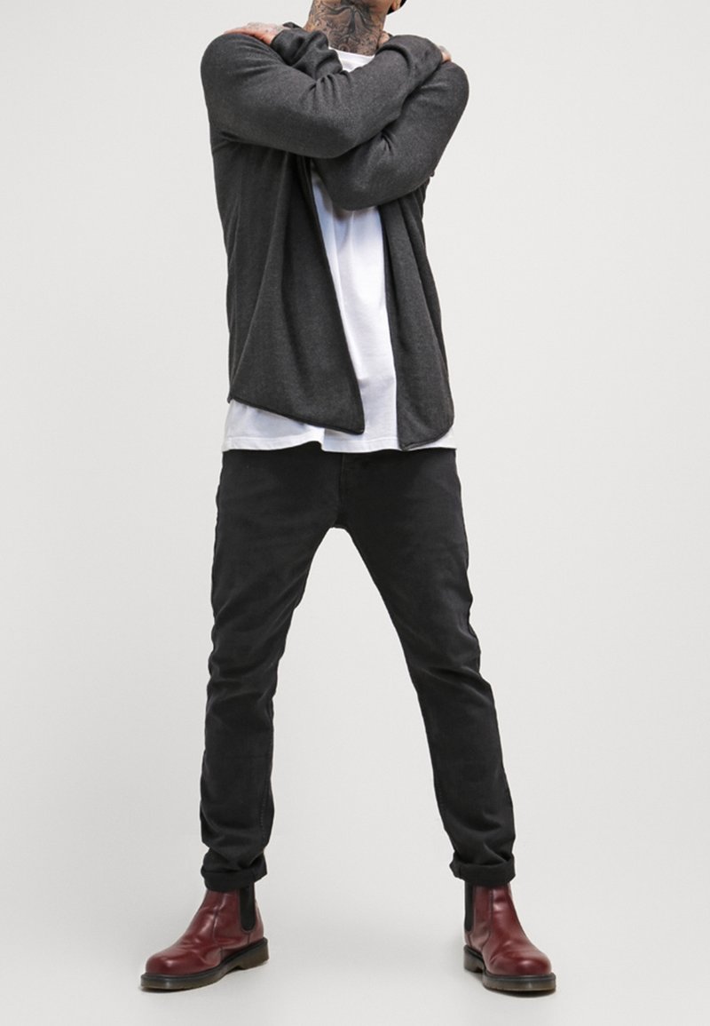 Szary kardigan na białej koszulce, w połączeniu z czarnymi spodniami i bordowymi butami typu chelsea. Gładka tkanina, luźny krój i podwinięte mankiety spodni.