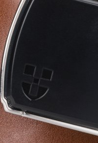 Czarna, błyszcząca odznaka z stylizowanym logo tarczy, otoczona srebrną obręczą, na teksturowanej brązowej powierzchni ze skóry.