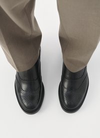 Sapatos brogue em couro preto com detalhes perfurados, parte superior lisa e bico redondo. Usados com calças castanhas claras, vistos de cima.