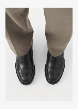 Schwarze Leder-Brogueschuhe mit durchbrochten Details, glattem Obermaterial und abgerundeter Zehenpartie. Getragen mit hellbraunen Hosen, von oben betrachtet.