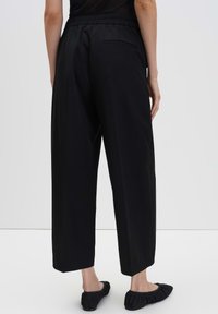 Achteraanzicht van een persoon die losse zwarte cropped broek met elastische tailleband en zwarte platte schoenen draagt op een witte achtergrond.