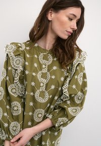 Kvinna med långt brunt hår som bär en olivgrön klänning med puffärmar och vita broderade cirkelmönster.