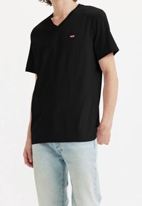 Camiseta de algodón negra de manga corta con escote en V, que presenta un pequeño logo rojo en el pecho y un corte relajado. Combinada con unos jeans de mezclilla azul claro.