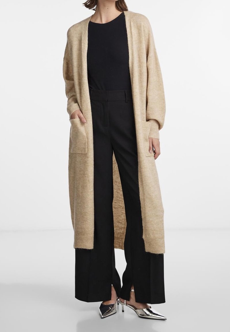 Personne portant un long cardigan beige, un haut noir, un pantalon noir à jambes larges et des escarpins argentés à bout pointu, debout devant un fond uni.