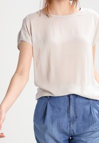 Krátký rukáv béžová blůza vyrobená z hladkého materiálu, s kulatým výstřihem a volným střihem, spárovaná s modrými džínovými kalhotami se záhyby.