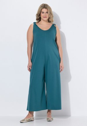 Kvinde med skulderlangt blond hår iført en turkis ærmeløs jumpsuit med brede ben og blomstrede slip-on sko, stående mod en hvid baggrund.