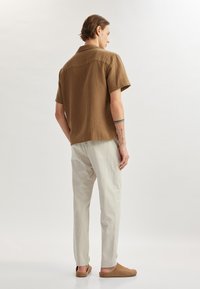Camisa marrón de manga corta en tejido texturizado, pantalones a rayas grises y zapatos sin cordones color marrón claro, vistos desde la parte de atrás.