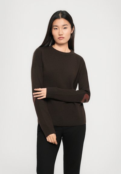 Young woman with long dark hair wearing a dark brown sweater with leather elbow patches and black pants, standing against a plain light background.